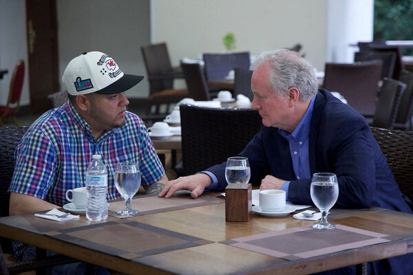 Two men sit at a square table talking in an otherwise empty cafeteria.